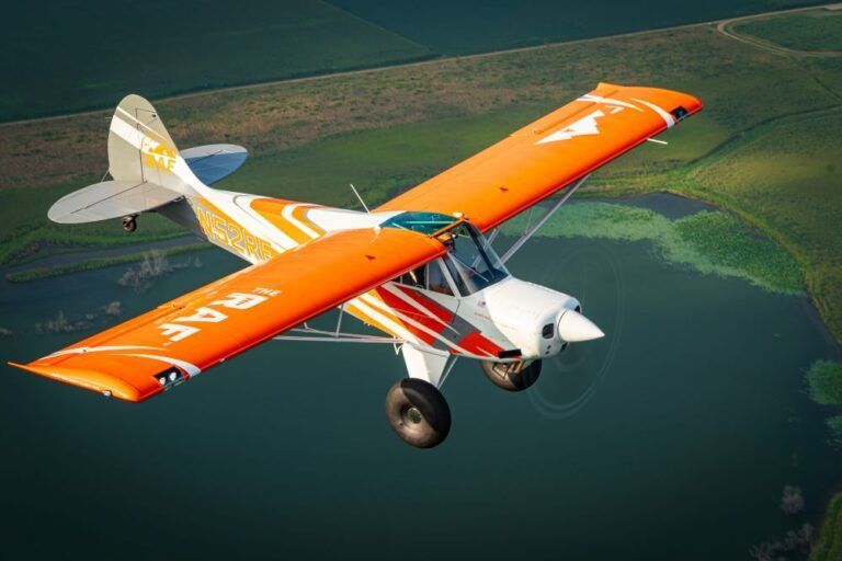 A small orange and white plane is flying over a body of water.