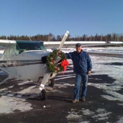 Man stands by a small airplane decorated with a wreath; snowy ground, sunny day.