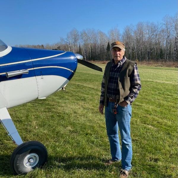 Man in jeans, plaid shirt, and vest, standing next to a blue and white airplane on a grassy airfield.