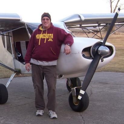 Man in maroon sweatshirt leans on a small airplane's nose, outdoors.