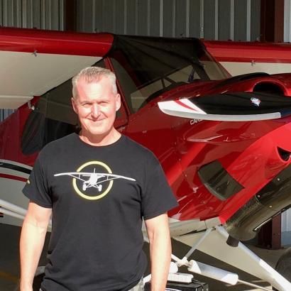 Man in black t-shirt smiles in front of a red and white airplane in a hangar.