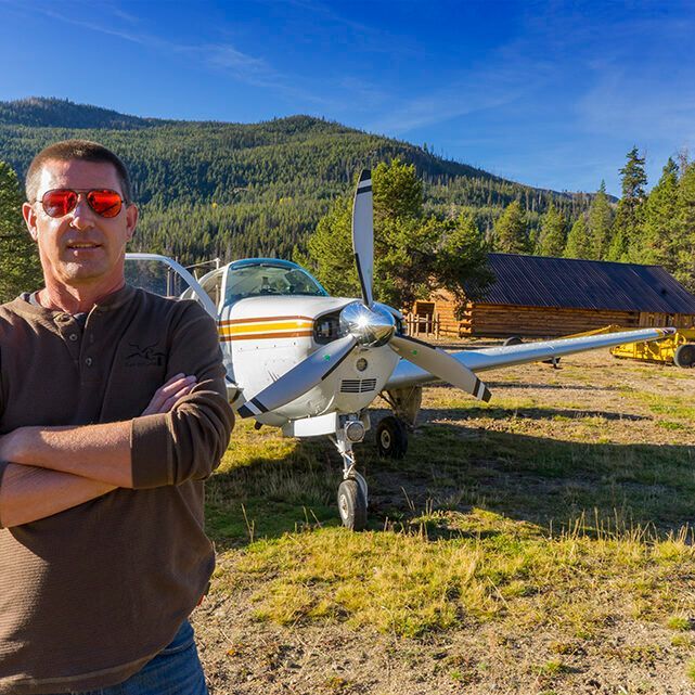 Man in sunglasses stands with arms crossed in front of small airplane and cabin in a mountain setting.