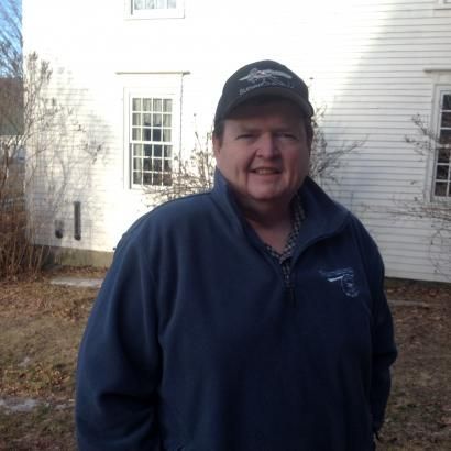 Man in blue sweatshirt and cap smiles in front of a white building.