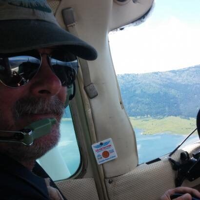 Man in sunglasses and cap piloting a small plane with a landscape visible outside the window.