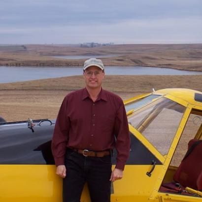 Man in maroon shirt stands near yellow airplane, smiling. Outdoors near a lake in a rural setting.
