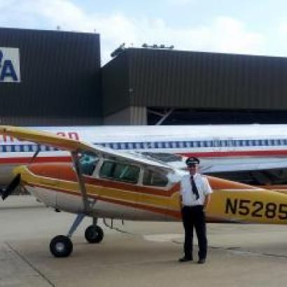 Pilot stands next to a small orange and yellow plane, with an American Airlines plane behind them in front of a hanger.
