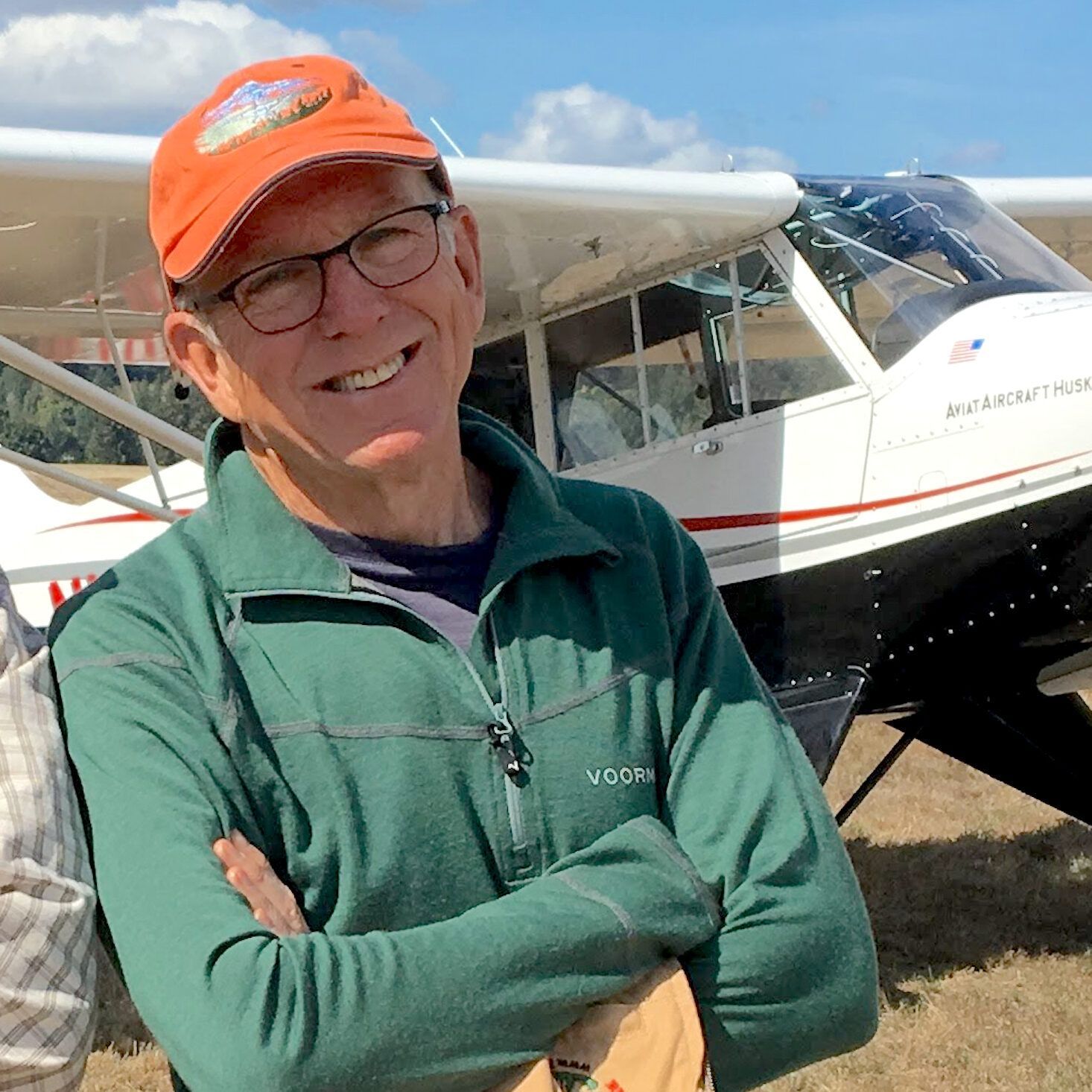 Smiling man in green jacket and orange cap with an airplane behind him.