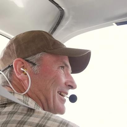 Man wearing a brown cap, headset, and checkered shirt, smiling inside an airplane cockpit.