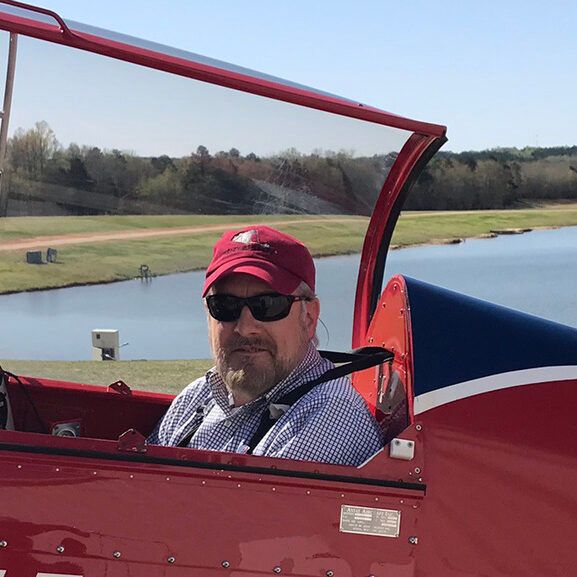 Man in red hat and sunglasses in cockpit of red and blue biplane, near a lake.