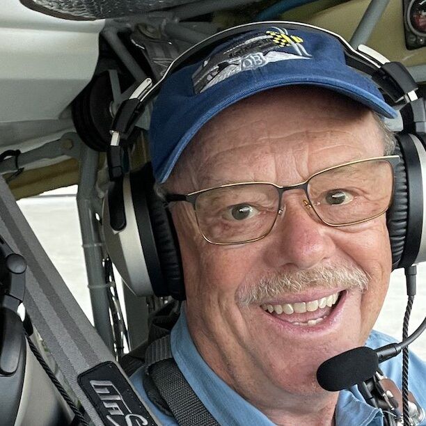 Smiling senior man in aircraft cockpit, wearing headset, glasses, and baseball cap.