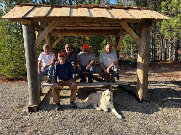 A group of men and a dog are sitting under a wooden shelter.