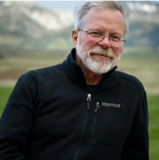 Man in cockpit of plane smiles, adjusting controls; wearing plaid shirt and jeans.