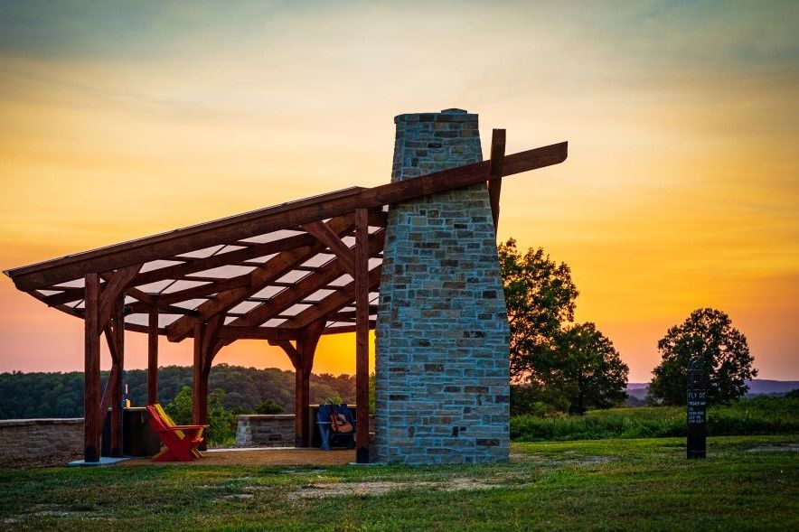A wooden pavilion with a stone chimney in the middle of a field at sunset.