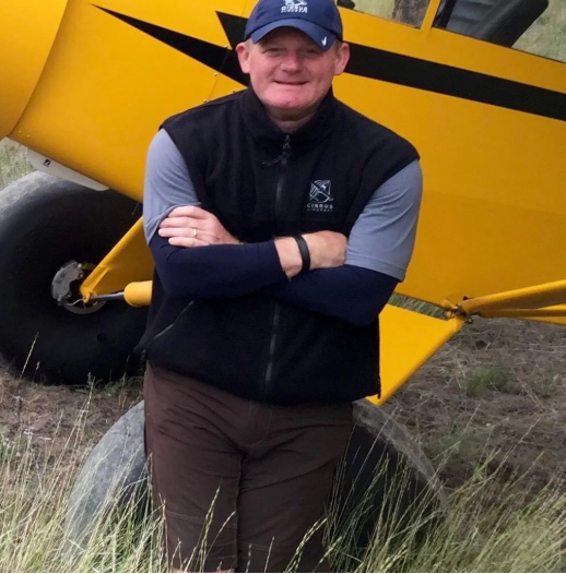 Man in cockpit of plane smiles, adjusting controls; wearing plaid shirt and jeans.