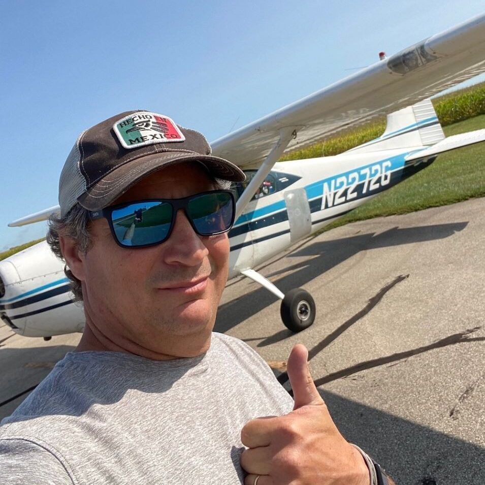 Man in sunglasses giving thumbs up in front of a small airplane with blue and white accents, sunny day.