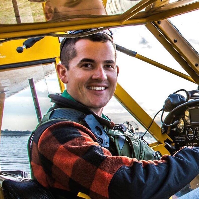 Man smiling, piloting a yellow plane with a lake in the background, wearing a plaid shirt and headset.
