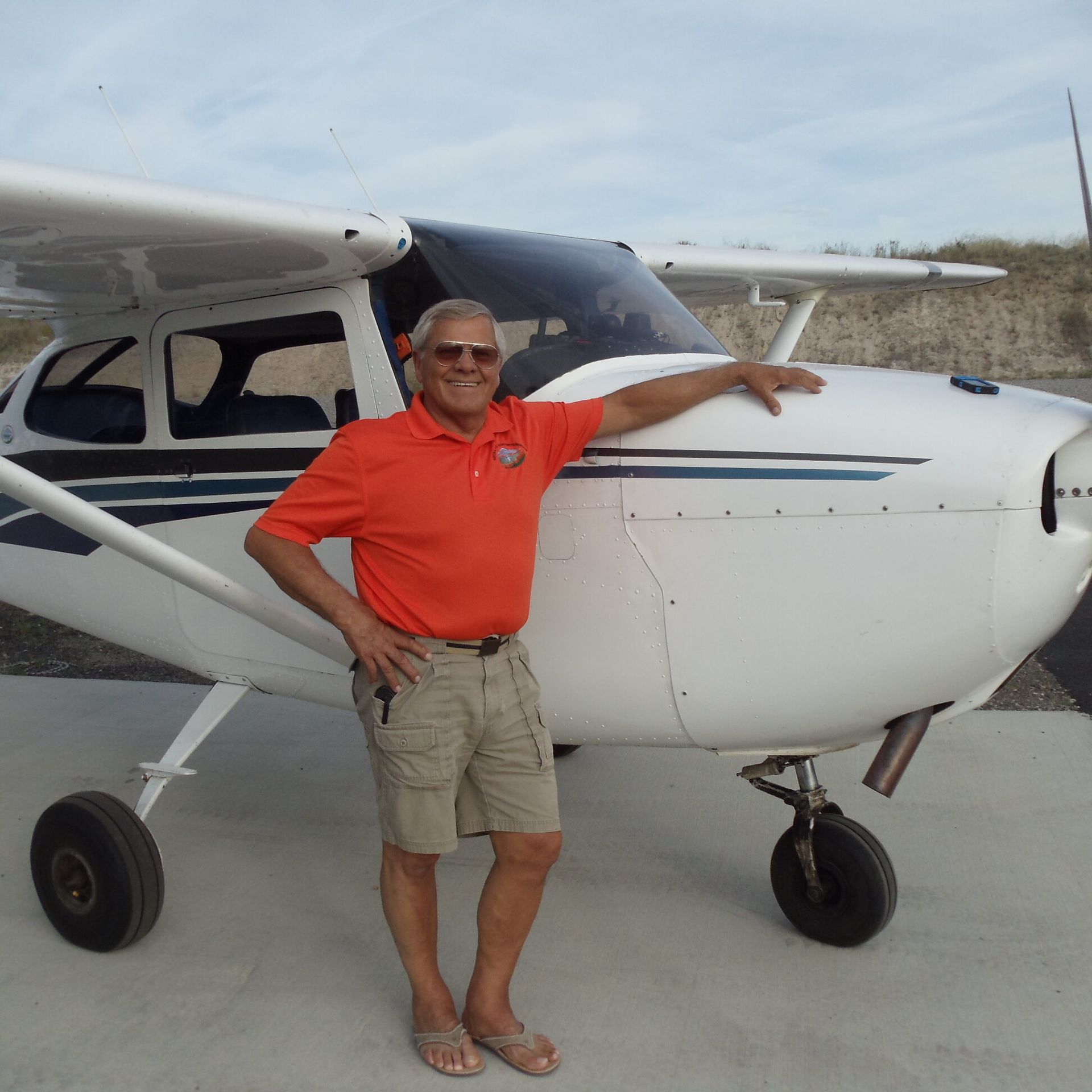 Man in orange shirt and khaki shorts poses beside a white airplane on a tarmac.