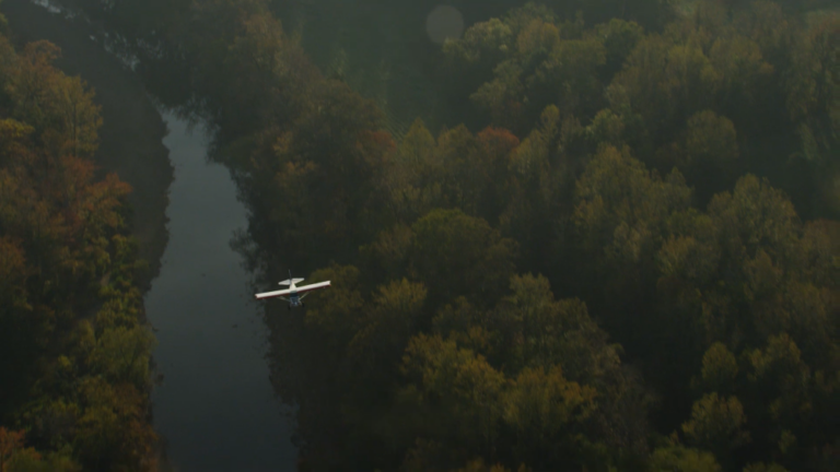 An aerial view of a small plane flying over a river surrounded by trees.