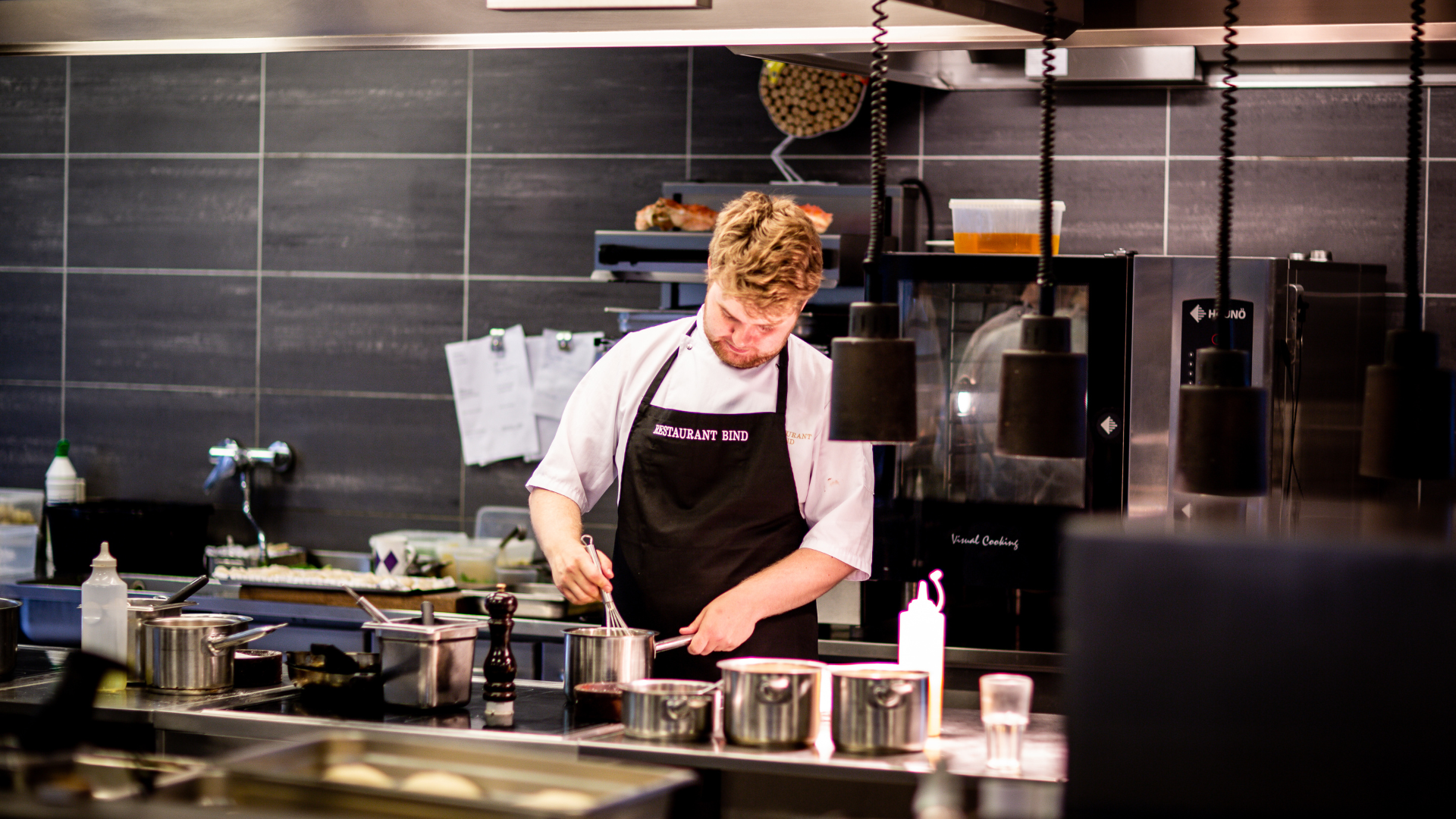 Chef in white shirt and black apron stirs pot in a commercial kitchen.