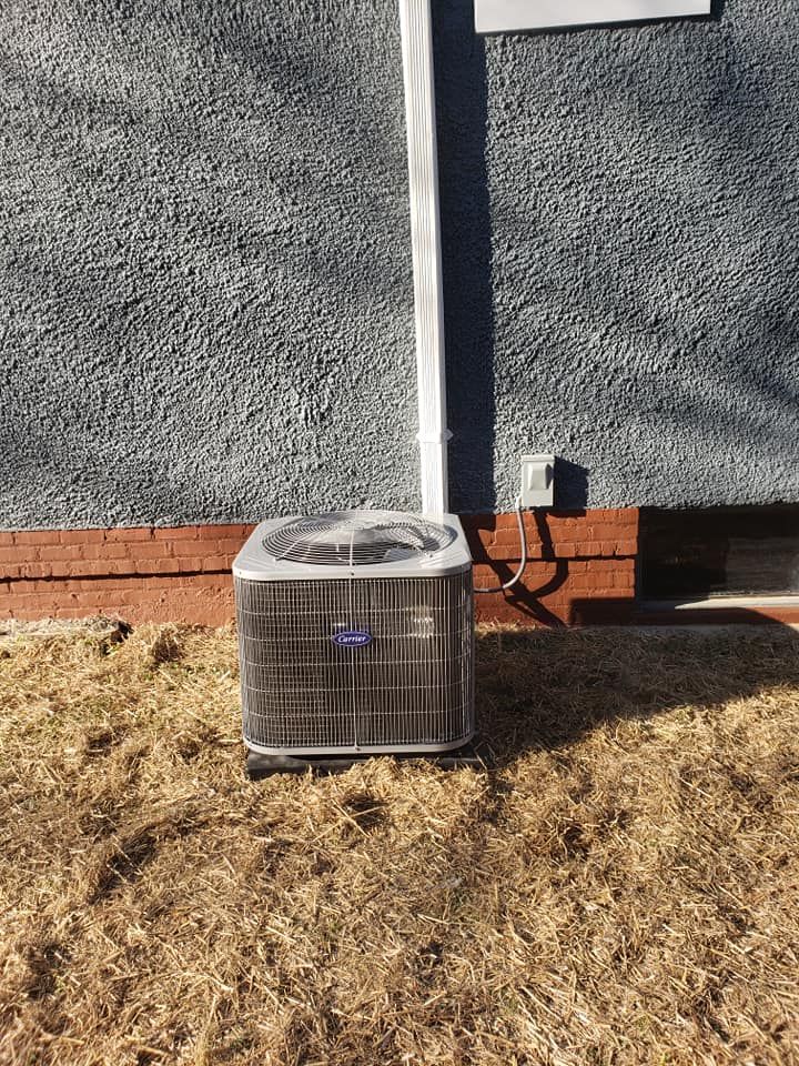 An air conditioning unit sits outside a building on dry grass. Brick foundation and gray stucco wall.