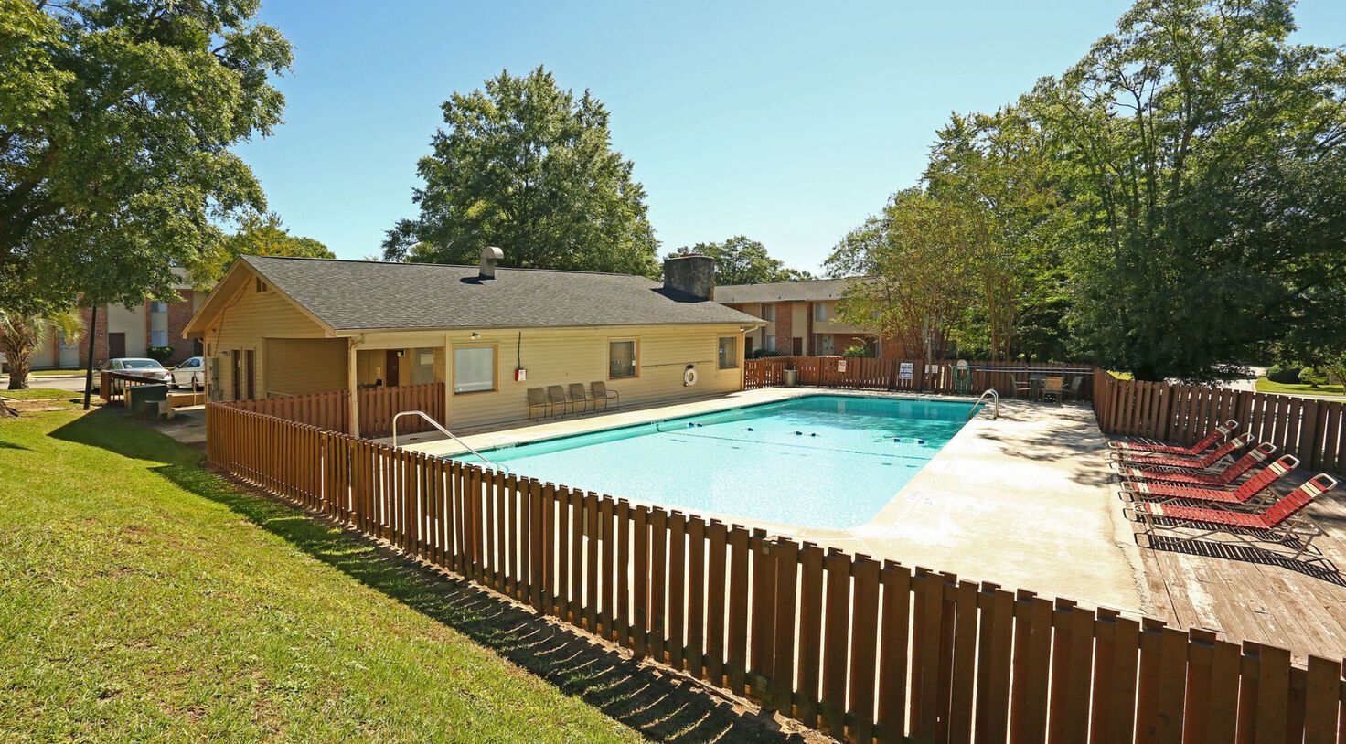 Swimming pool with a wooden fence, a building, and lounge chairs on a sunny day.