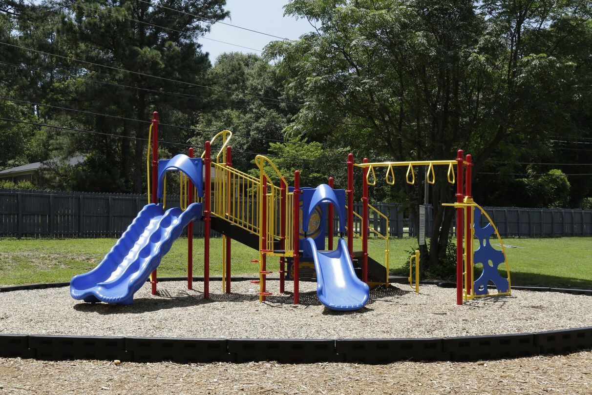 Playground with blue slides, yellow and red climbing structure, on wood chips, surrounded by trees.