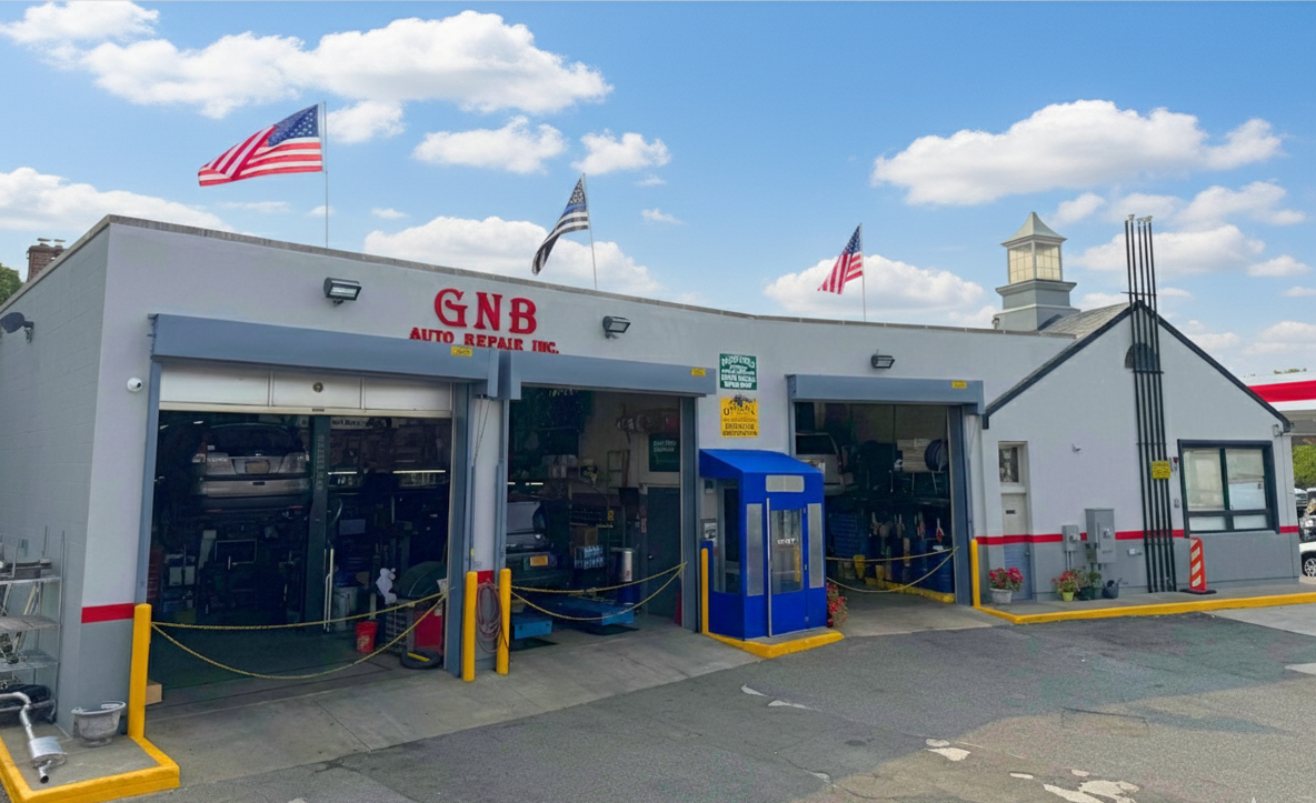 Exterior of a grey GNB auto repair shop with garage doors open, three American flags and a small lighthouse-like structure.