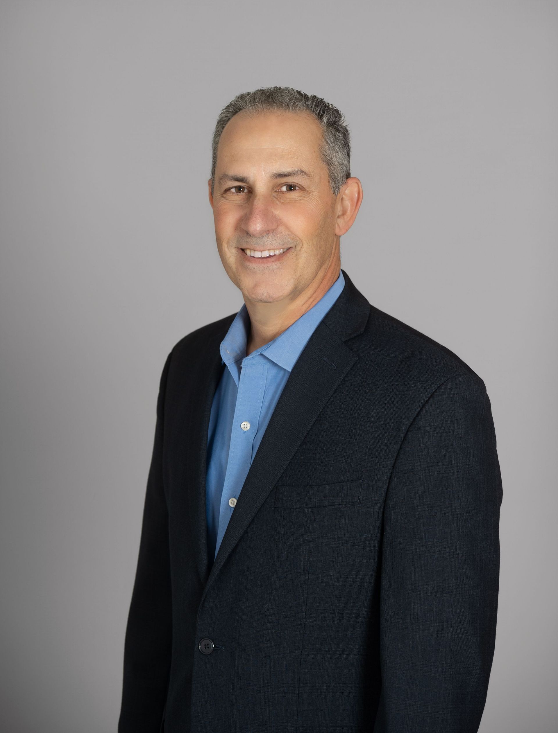 Man in a blue shirt and navy blazer smiling at the camera against a gray background.