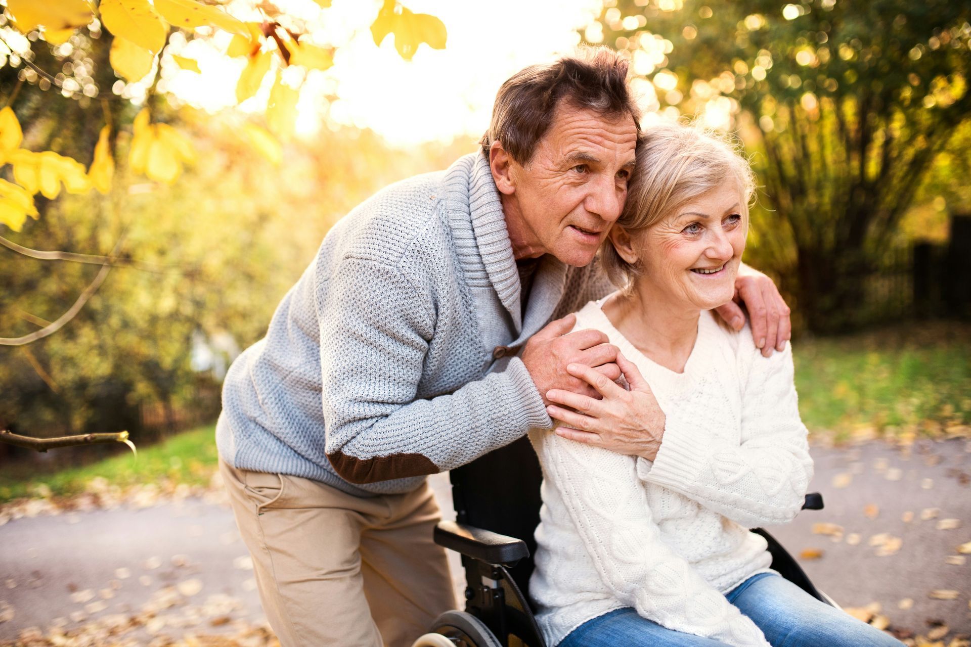 Man with arm around woman in wheelchair, smiling, outdoors in autumn.