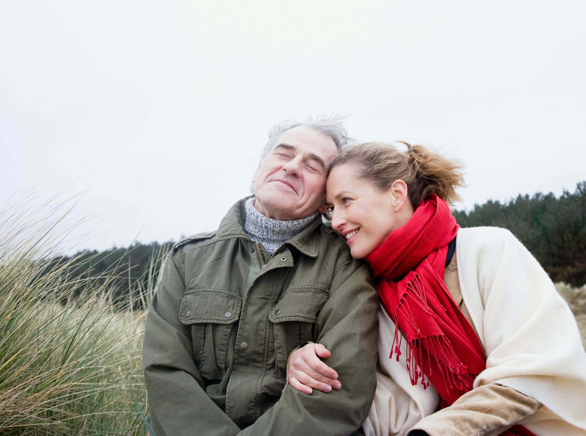 Couple embraces outdoors; man smiles, woman leans on him, red scarf.