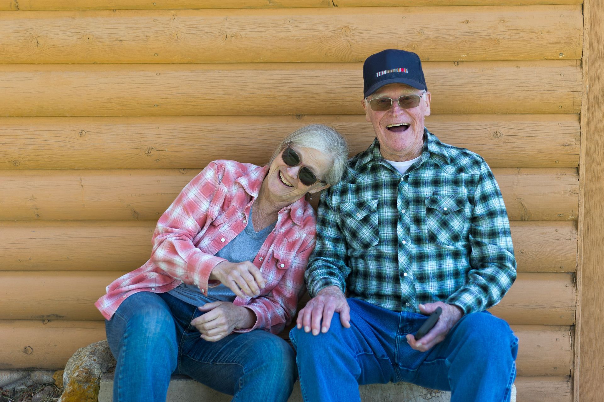 Couple sitting and smiling in front of a wood-paneled wall, wearing casual clothes and sunglasses.
