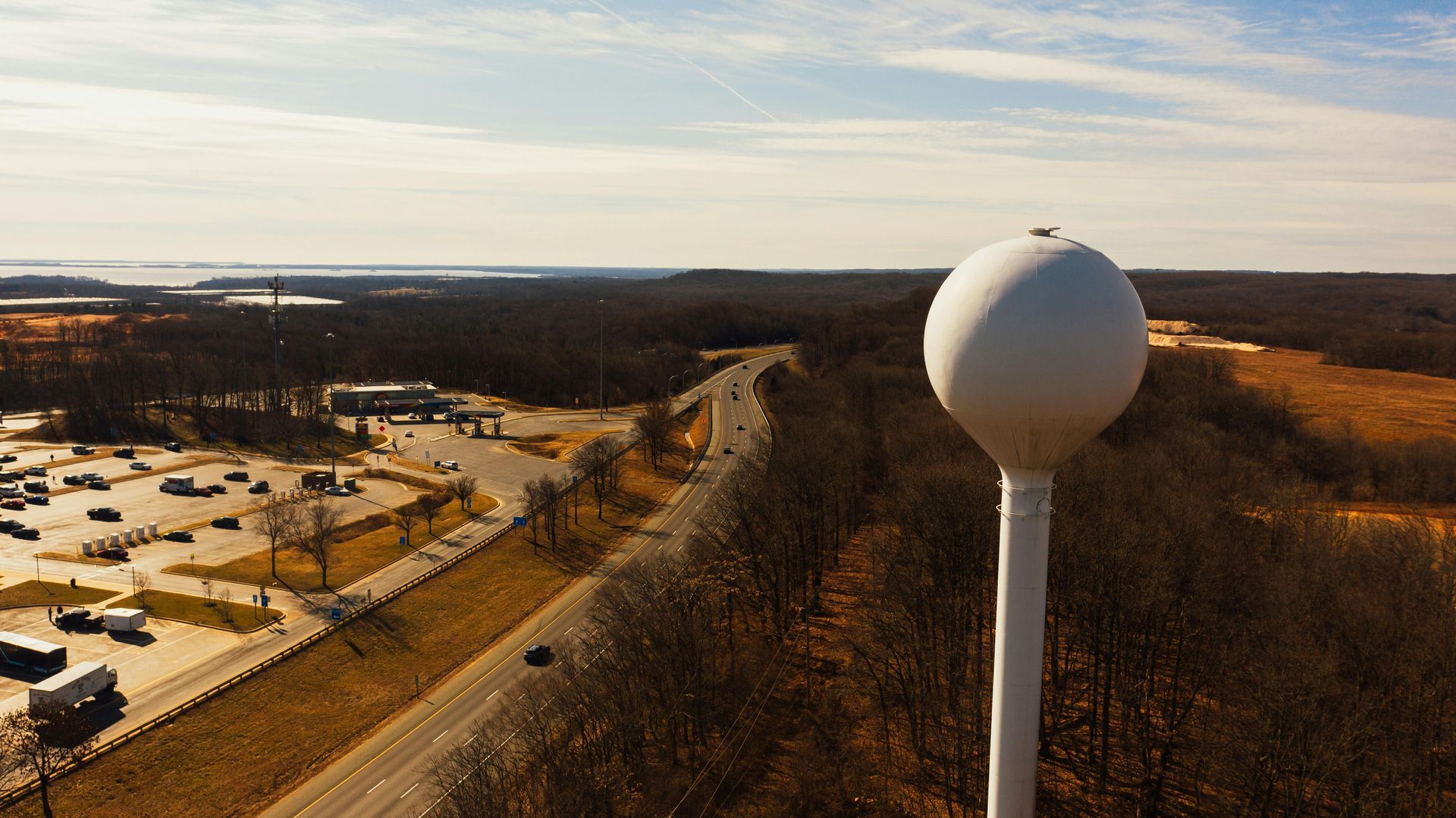 Water tower on a ridge overlooking a highway, parking lot, and forest under a partly cloudy sky.