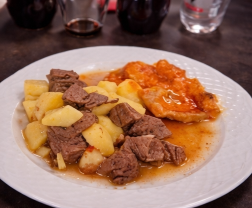 Un piatto bianco contenente spezzatino di manzo con patate e un contorno di verdure saltate di colore arancione in una salsa saporita.