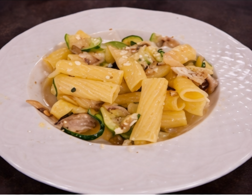 Una ciotola bianca piena di rigatoni conditi con zucchine e funghi saltati in padella, leggermente cosparsa di formaggio.
