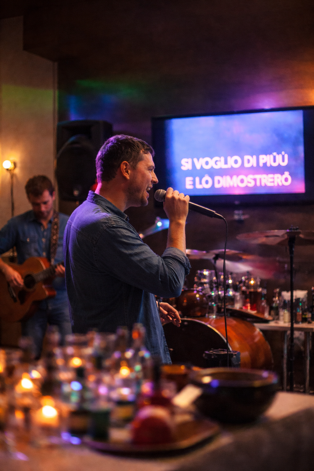 Un uomo canta in un microfono sul palco, con un chitarrista sullo sfondo e il testo della canzone visualizzato su uno schermo.