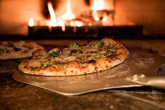 Una pizza con condimenti verdi su una pala di metallo, viene estratta da un forno a legna con fiamme visibili sullo sfondo.