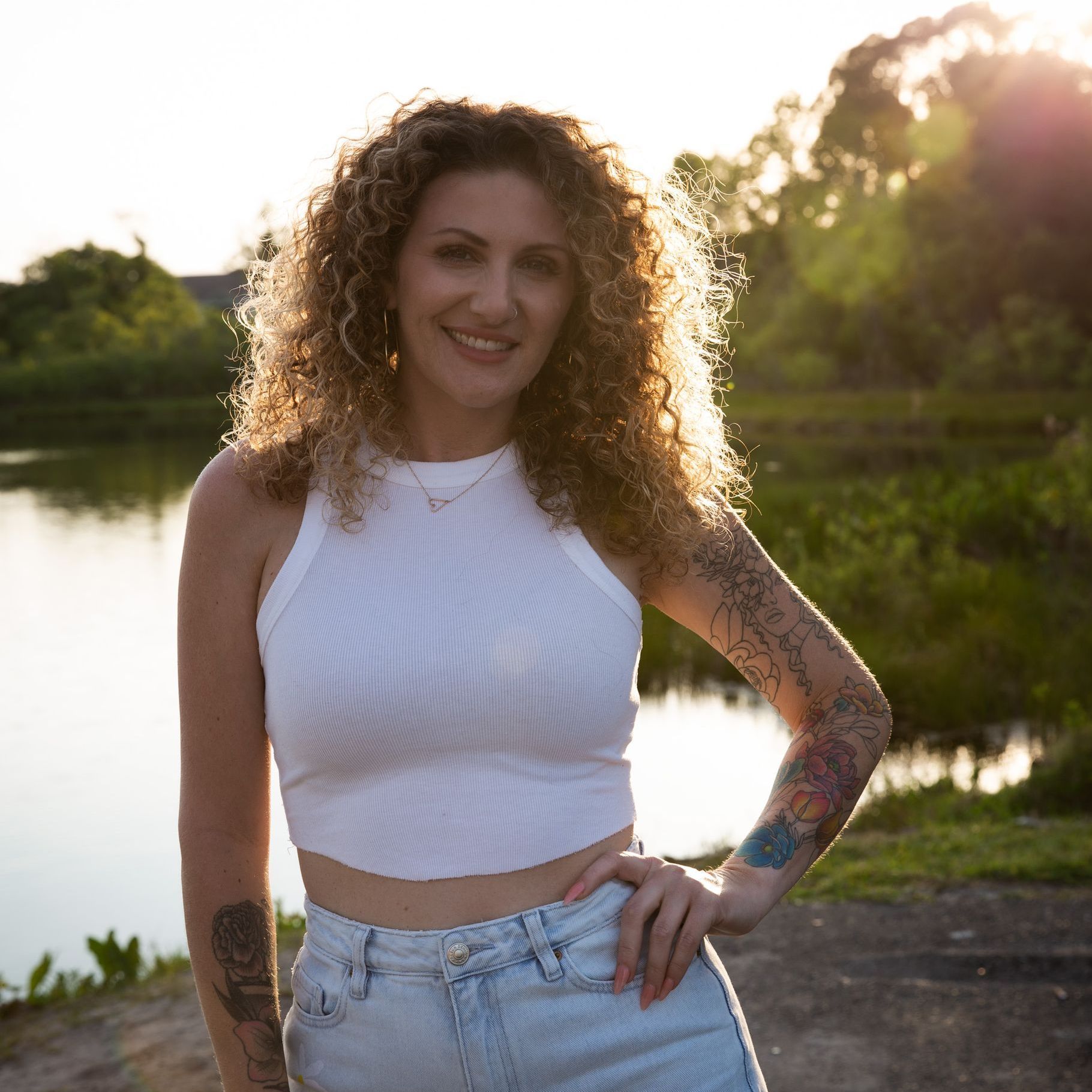 A woman with curly hair and tattoos is standing in front of a body of water.