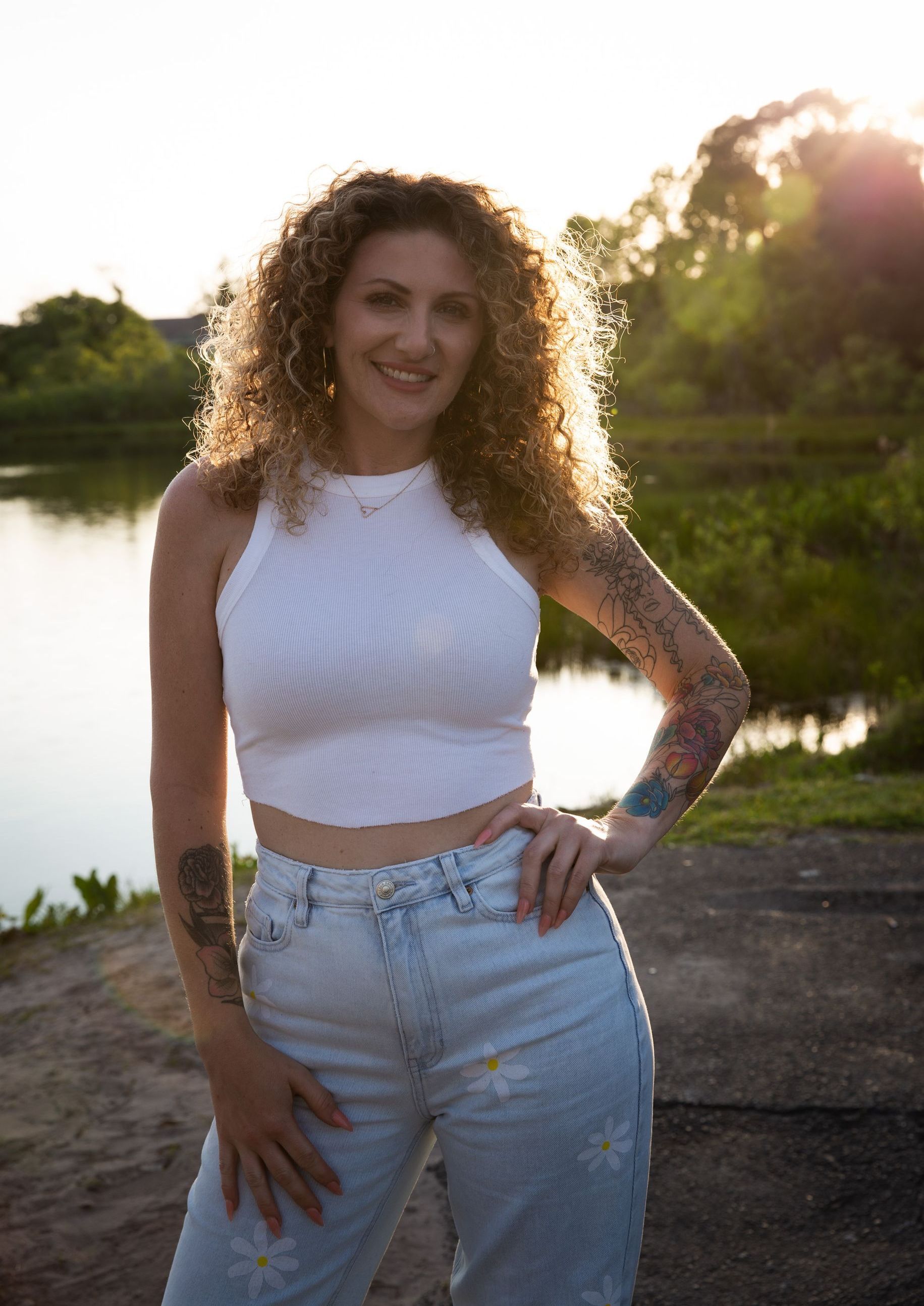 A woman in a white crop top and jeans is standing in front of a lake.