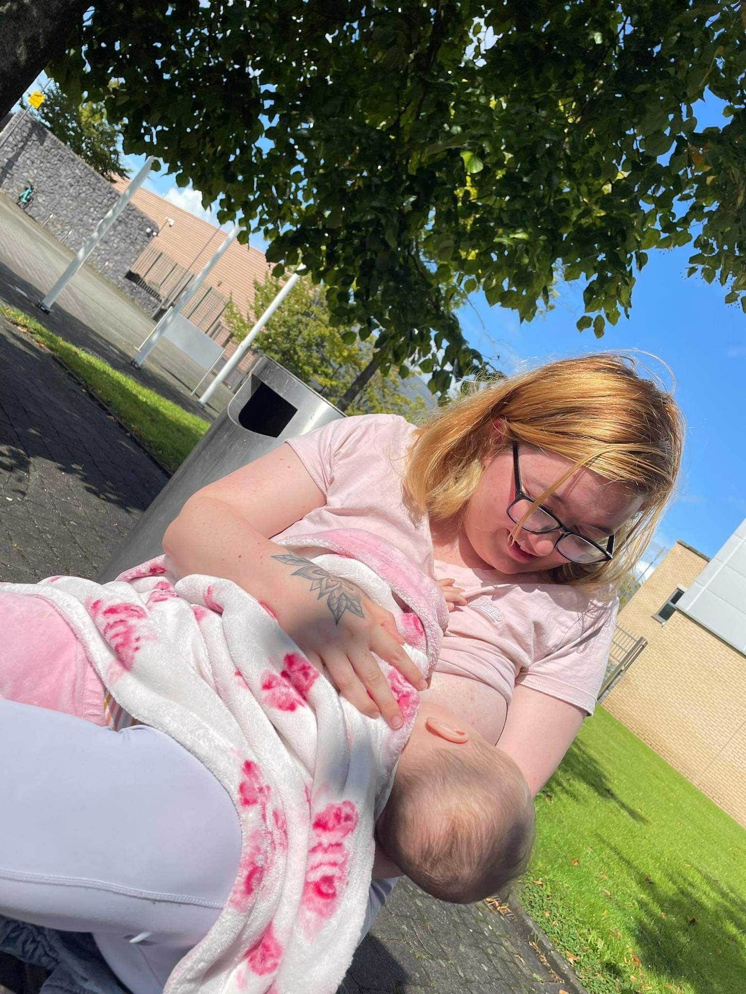 mother with blond hair and a pink t-shirt sitting under a tree on a sunny day, breastfeeding her baby