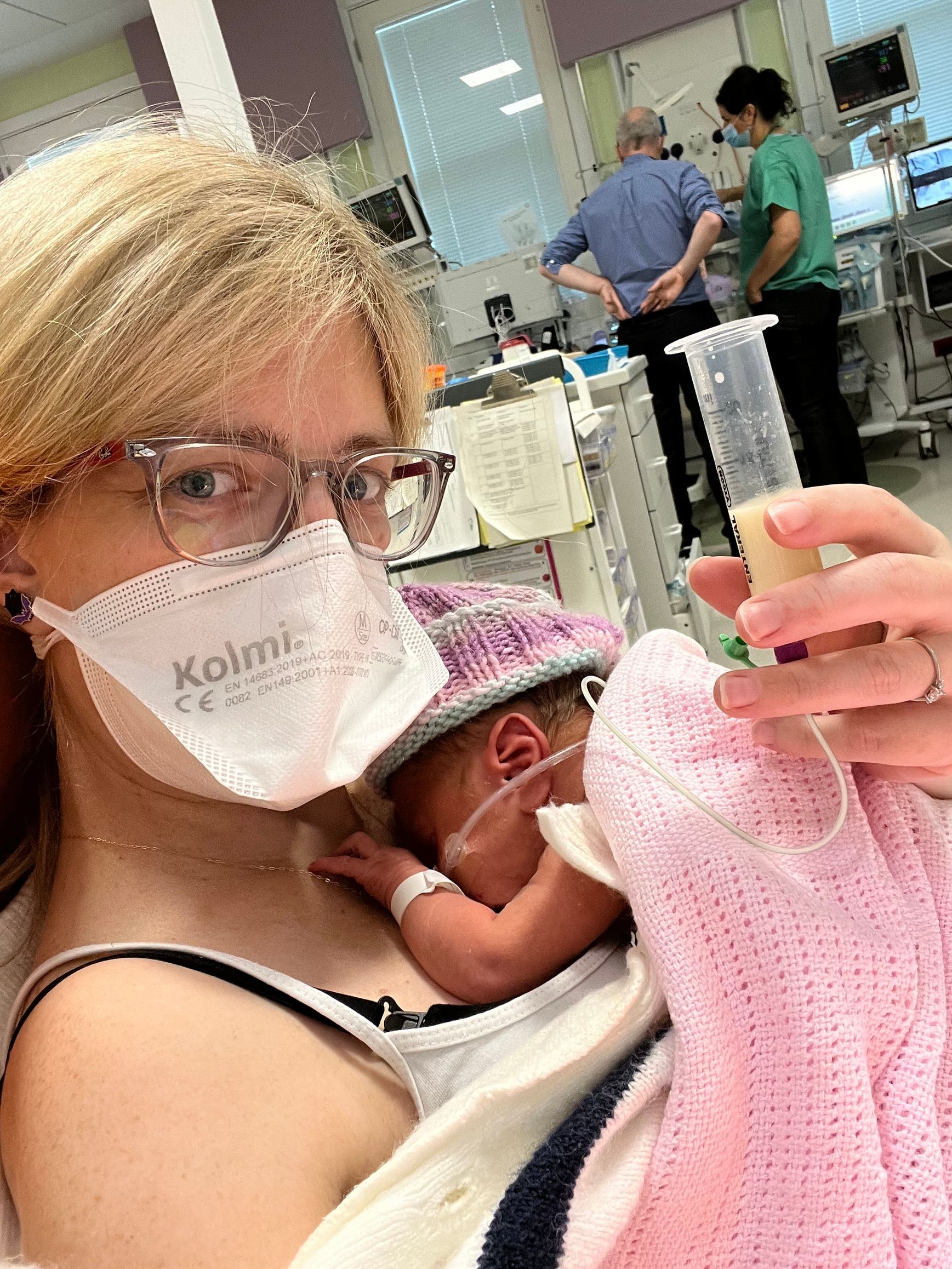 mother with surgical mask, with her baby on her chest skin to skin, feeding her baby with a purple knitted hat,  with breastmilk via a feeding tube.
