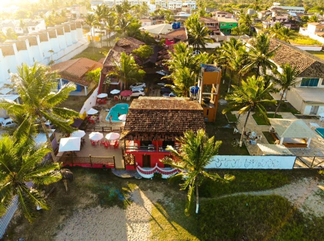 Vista aérea de um edifício com telhado vermelho perto de uma praia, com piscina, palmeiras e outros edifícios.
