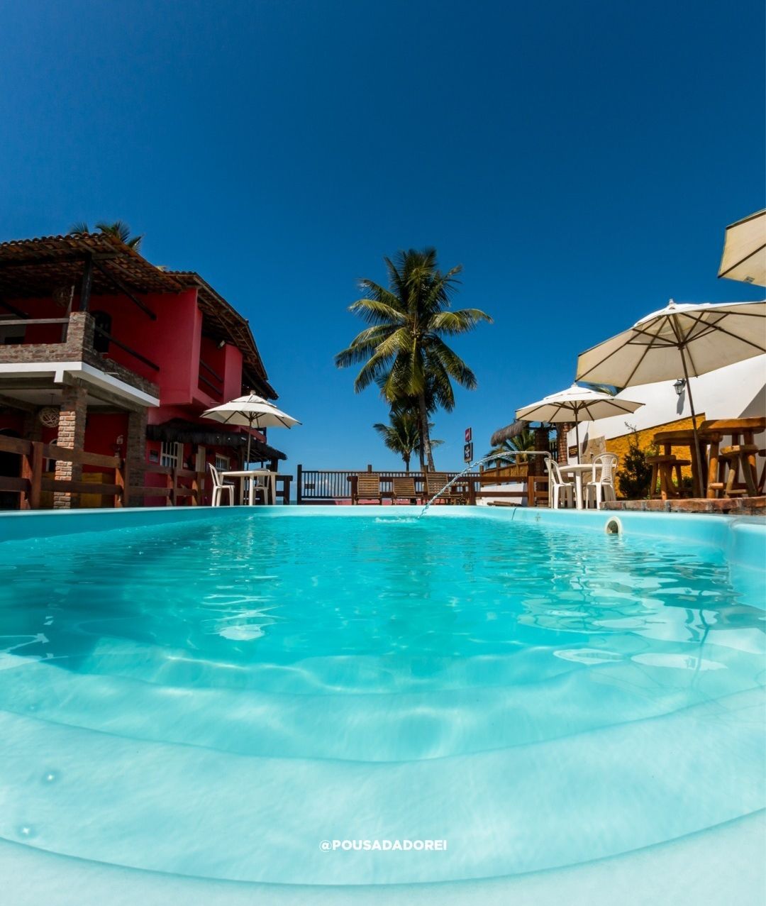 Piscina com água cristalina perto de prédios e palmeiras sob um céu azul brilhante.
