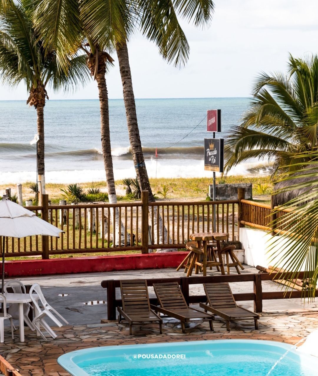 Piscina, praia e palmeiras. Deck de madeira marrom com espreguiçadeiras, oceano ao fundo.