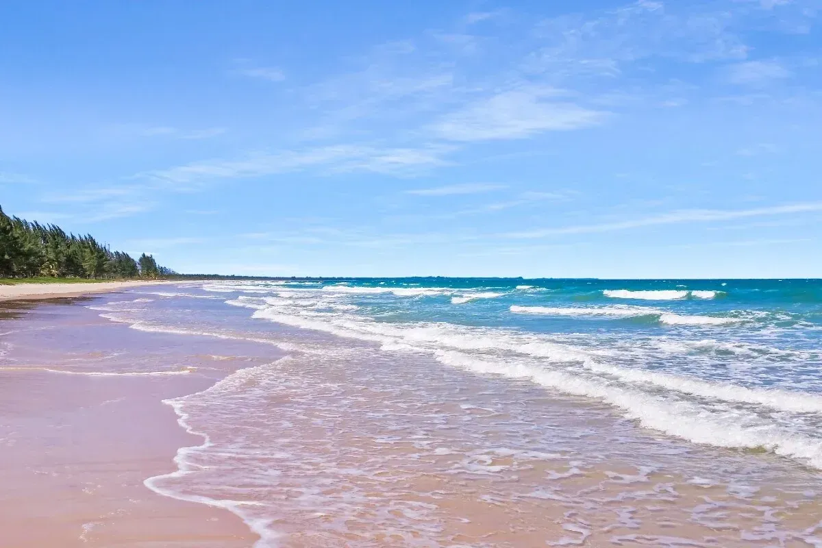 Praia de areia com oceano e céu azuis; ondas suaves quebram na costa.