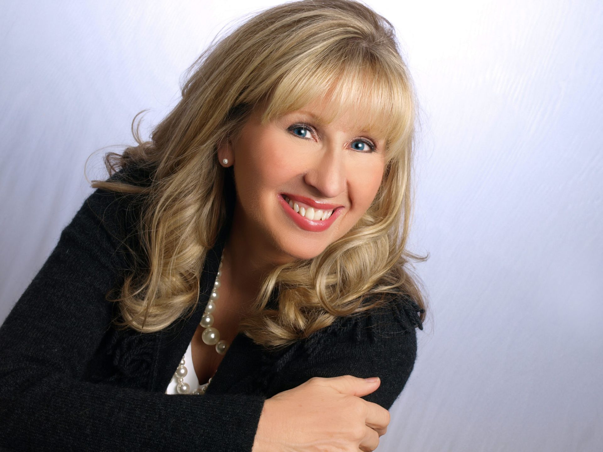 A smiling woman with blonde hair, wearing a black blazer and a pearl necklace, poses against a white background.