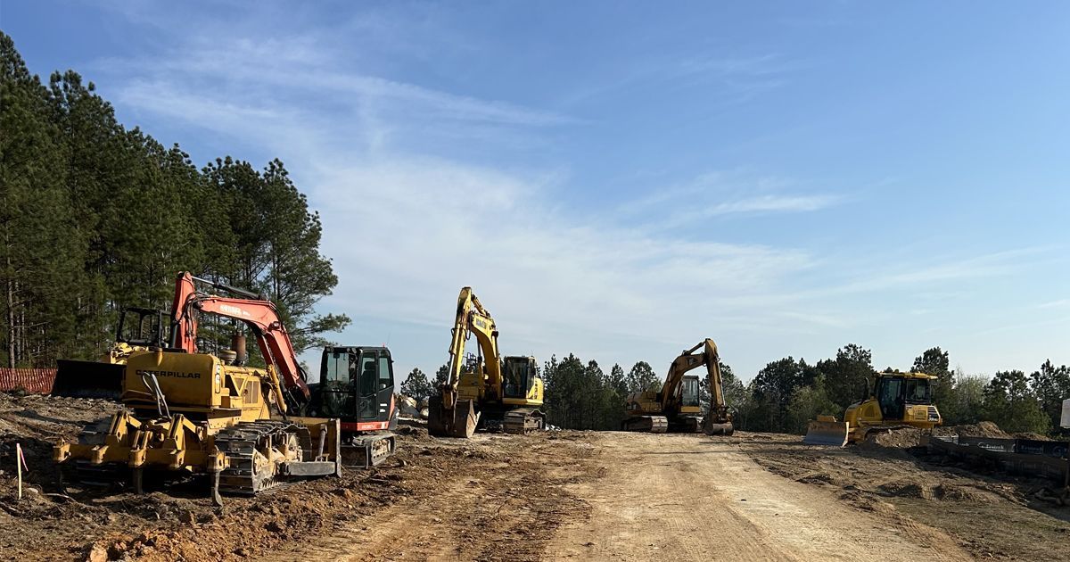 Four construction excavators parked on a dirt path at a clearing with a treeline under a clear blue sky.