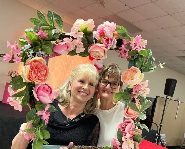 Two smiling individuals pose together holding a decorative floral heart-shaped frame in an indoor setting.