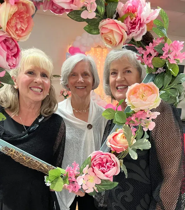 Three smiling people pose together behind a large, circular, flower-adorned picture frame.