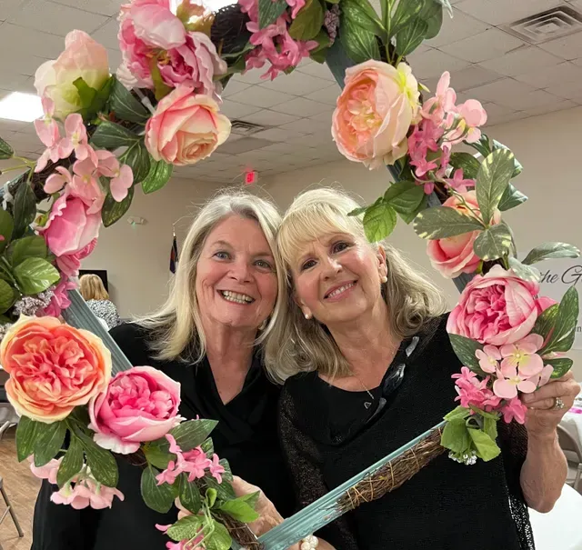 Two smiling individuals pose behind a large oval frame decorated with pink and peach artificial flowers.