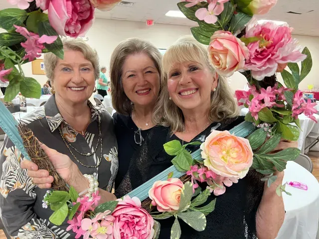 Three smiling individuals pose together behind a decorative floral frame adorned with pink flowers and greenery.